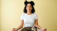 Young woman with curly dark hair and medium skin tone meditating with tabby cat on lap against yellow background for wellness and pet lifestyle content.