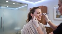 Professional makeup artist applying eyebrow makeup to smiling Caucasian woman client in modern beauty salon with soft lighting and contemporary interior.