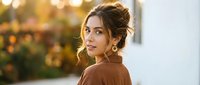 Young woman with elegant updo hairstyle looking over shoulder in warm golden hour light wearing brown sweater for lifestyle and beauty content.