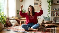 Young woman celebrating success with raised fists in cozy boho living room with plants and natural light streaming through windows.