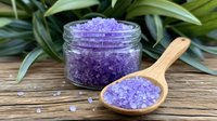 Purple bath salt crystals in glass jar and wooden spoon on rustic wood surface with green tropical leaves, spa wellness concept for aromatherapy and relaxation.