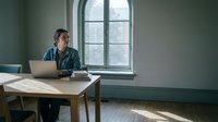Young Caucasian man working on laptop at wooden desk by arched window in minimalist green room with natural light for remote work concepts.