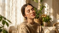 Middle-aged woman with brown hair wearing beige knit sweater sitting indoors near potted plants and lace curtains, looking thoughtful in natural daylight for lifestyle and wellness content.