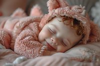 Sleeping baby with red hair wearing soft pink bear costume rests peacefully on patterned bedding, capturing innocent childhood moments.