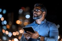 Middle Eastern businessman using digital tablet at night with urban bokeh lights background, wearing glasses and casual business attire, looking upward thoughtfully.