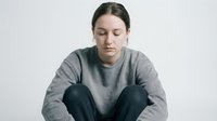 Young Caucasian woman in gray sweatshirt sitting cross-legged with eyes closed in peaceful meditation pose against neutral background for wellness content.
