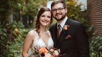 Young couple in wedding attire smiling outdoors, bride in lace white dress and groom in dark suit with burgundy tie holding autumn-themed floral bouquet against natural foliage.