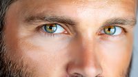 Close up portrait of male face with striking multicolored eyes and intense gaze against dark background, showing partial features in dramatic lighting.