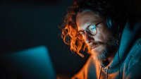 Focused bearded man with glasses and headphones working late at computer in dark room with colorful ambient lighting for gaming or creative projects.