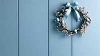 Delicate wreath with white berries and blue satin bow hanging on light blue wooden planked wall, creating minimalist holiday decoration style.