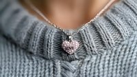 Delicate silver necklace with pink love heart pendant resting on soft gray knitted sweater, creating romantic Valentine's day or anniversary jewelry composition.