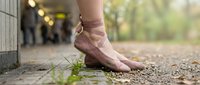 Young female ballet dancer in pointe shoes standing on outdoor ground surface near building wall, practicing classical dance technique in natural daylight setting.