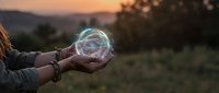 Hands holding glowing transparent sphere with magical light effects against sunset landscape background for fantasy and mystical concepts.