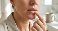 Middle-aged Caucasian woman touching lips with finger in contemplative gesture, showing concern or worry in bright kitchen setting for wellness concepts.