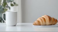 Fresh baked croissant and white ceramic coffee mug on minimal white table setting with soft natural lighting and blurred green plant background.