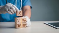 Healthcare professional in blue scrubs stacking wooden blocks with medical icons including stethoscope and heart symbols on white desk.