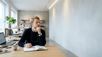 Mature man with light brown hair and fair complexion, wearing dark blazer and headphones around neck, sitting at wooden desk in modern office space with laptop and notebook, working in contemporary workplace environment.