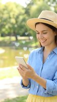 Young woman in straw hat using smartphone with heart icons floating from screen, smiling while texting in sunny park setting for social media and dating apps.