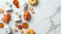 Halloween decorations with orange skull, white ghosts, pumpkins and autumn flowers arranged on marble background for seasonal party planning.