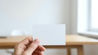 Cropped hand holding blank white business card or invitation mockup against wooden table and bright window background for branding and marketing.