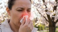 Middle-aged Caucasian woman suffering from seasonal allergies blowing nose with tissue outdoors during spring cherry blossom season for health and wellness concepts.