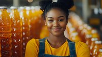 Young African American woman in yellow top and overalls smiling warmly at camera with rows of orange juice bottles creating vibrant background blur in retail setting.