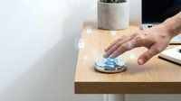 Wireless charging pad on wooden desk with hand reaching to place smartphone, modern office workspace with plant and laptop for technology concepts.