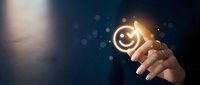 Businessman hand holding glowing smiley face icon against dark blue background with bokeh lights, representing customer satisfaction, positive feedback, and service quality for business concepts.