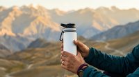 Hiker holding white water bottle with mountain range in background during golden hour, outdoor adventure and hydration concept.