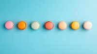 Colorful macarons arranged in straight line on turquoise background. Sweet French dessert cookies in pastel pink, orange, cream, coral, beige and yellow colors.