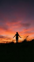 Woman silhouette with open arms against dramatic sunset sky with orange and purple clouds over grassy field for freedom and wellness concepts.