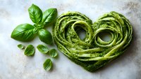 Fresh green pasta shaped as heart with homemade basil pesto sauce and aromatic herbs on light marble background, creative food styling for Valentine's day menu.