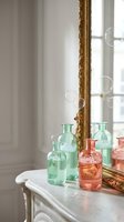 Colorful glass bottles in green and coral pink arranged on white vintage table with ornate gold mirror and floating soap bubbles in elegant interior.