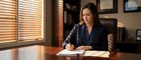 Professional businesswoman writing notes at wooden desk in modern office with warm sunlight streaming through blinds creating dramatic shadows.