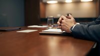 Professional businessman hands clasped on wooden conference table during corporate meeting with documents and water glass in modern boardroom.