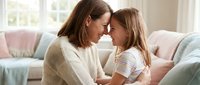 Loving mother and daughter touching foreheads and smiling together on sofa at home, sharing a tender bonding moment in bright living room.