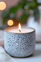 Lit aromatic candle in speckled ceramic holder with warm glow against soft bokeh background creates cozy atmosphere for wellness and meditation spaces.