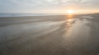 Serene coastal landscape at sunrise with golden sunlight reflecting across wet sand and shallow tidal waters, creating rippled patterns on the beach shore.