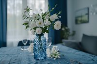White delicate flowers in blue crystal vase on dining table with textured tablecloth, soft natural window light creates moody atmosphere in modern interior.