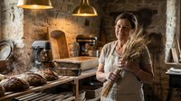 Elderly Caucasian female baker holding wheat stalks in rustic stone bakery with fresh bread loaves and traditional baking equipment for artisan food business.
