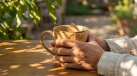 Elderly hands holding warm coffee mug in golden morning sunlight outdoors. Senior person enjoying peaceful breakfast moment in garden setting.