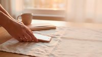 Hand touching smartphone on wooden desk with coffee cup and notebook in warm morning sunlight for business communication and productivity workspace.