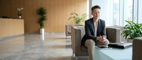 Professional businesswoman in suit sitting in modern office lobby with plants and natural lighting for corporate communications.