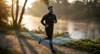 Athletic man jogging along misty river path during golden hour sunrise. Morning fitness routine in peaceful natural setting with warm sunlight filtering through trees.