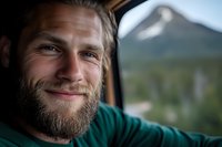 Smiling bearded Caucasian man in green shirt taking selfie inside car with mountain landscape visible through window. Natural lighting creates warm atmospheric portrait.