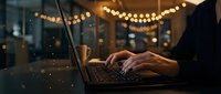Professional working on laptop in cozy evening cafe with warm string lights creating productive atmosphere for remote work and digital nomad lifestyle.