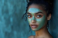 Young African American woman with curly hair wearing turquoise clay face mask for skincare routine and spa treatment against blue background.