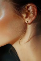 Close-up view of woman's ear with small gold hoop earring, showing delicate skin texture and wisps of hair in warm natural lighting against dark clothing.