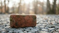 Old weathered red brick lying on gravel stones with blurred autumn forest background. Rustic construction material concept for renovation projects.