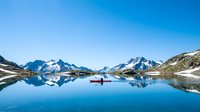 Serene alpine lake with snow-capped mountain peaks reflected in calm blue water, featuring a lone red boat, ideal for travel and nature backgrounds.